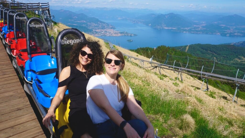 Ragazze sorridenti alla partenza di Alpyland, le montagne russe in vetta al monte mottarone da cui si gode di una vista mozzafiato sui sette laghi, tra cui il lago maggiore alle loro spalle.